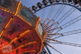 Impression von einem Jahrmarkt. Im Vordergrund ein Teil des mit nostalgischen Bildern und Beleuchtung verzierten Oberteils eines Kettenkarussells. In einer glatten Fläche spiegelt sich noch die Helligkeit des Widerscheins der untergehenden Sonne. im Hintergrund steht ein Riesenrad, auch von ihm ist nicht alles zu sehen, auch hier ist die Beleuchtung bereits angeschaltet. Durch die filigrane Konstruktion ist der Abendhimmel zu sehen, der noch hell und blau ist, die Sonne geht im Moment erst unter.