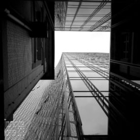 schwarzweiß-Aufnahme. Ein altes 2-stöckiges Haus und direkt daneben ein modernes Hochhaus mit Glasfassade. Perspektive von unten senkrecht nach oben, der Fluchtlinie der Fassaden folgend zum weiß überstrahlten Himmel.