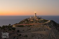 Landschaftsaufnahme: Ein Leuchtturm auf einem kargen, felsigen Hügel am Meer. vom Leuchtturm herunter windet sich in Serpentinen eine Straße, auf der die Lichter von Autos zu sehen sind, durch Langzeitbelichtung zu einem Lichtstreifen verwischt.