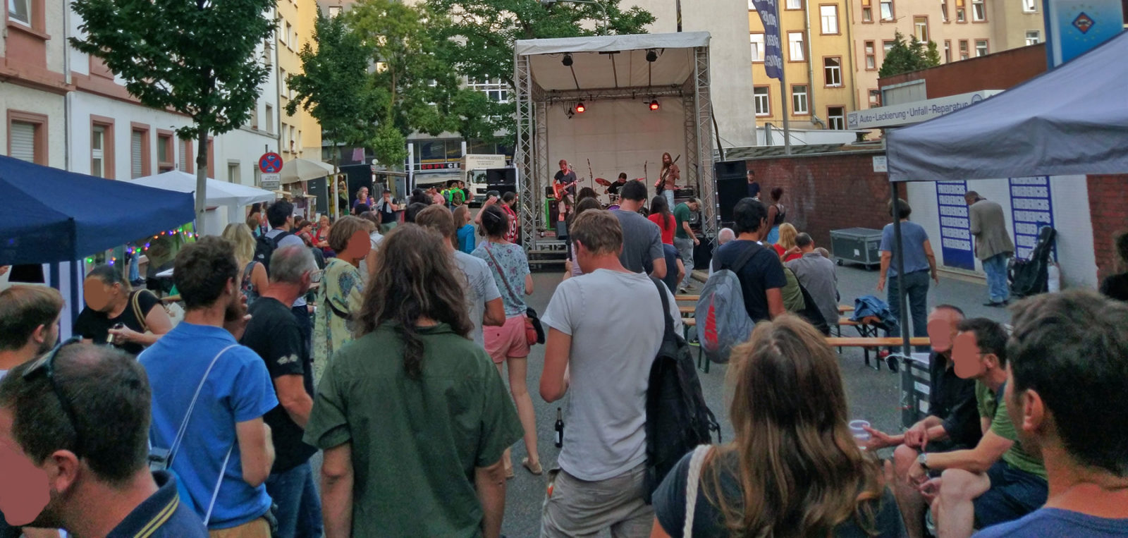 Totale über das Straßenfest. Im Vordergrund etliche Besucher von hinten, im Hintergrund vor den Besuchern die Bühne mit Hazard Dawn