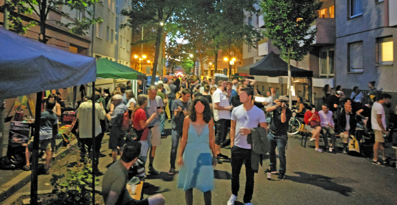 Totale über das Straßenfest. Es ist bereits dämmrig, die Straßenbeleuchtung ist an. Die Besucher scharen sich um Stände oder schauen zur Bühne, die Straße ist voll