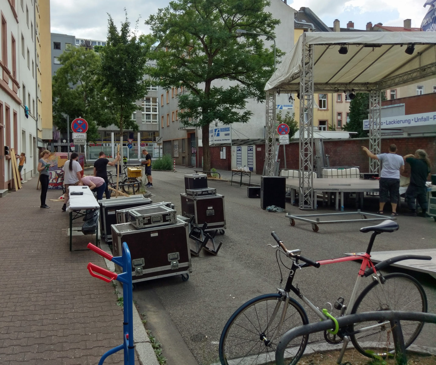 Aufbau des Straßenfests: Auf der Straße eine halbfertige Bühne, Verpackungskoffer von Bühnenequipment, Tische, eine Sackkarre und Gruppen von arbeitenden Leuten