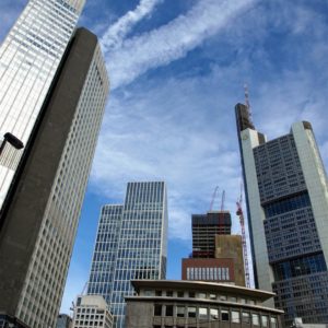 Stadtansicht von Frankfurt. Bild im Hochformat unter blauem Himmel mit Kondensstreifen von Flugzeugen, im Vordergrund Wolkenkratzer, Hoch- und Bürohäuser des Bankenviertels, von den 50er Jahren bis gerade im Bau