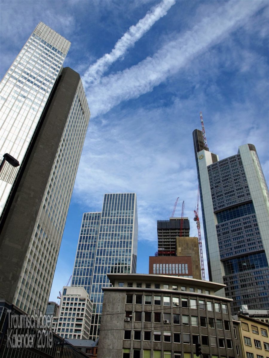 Stadtansicht von Frankfurt. Bild im Hochformat unter blauem Himmel mit Kondensstreifen von Flugzeugen, im Vordergrund Wolkenkratzer, Hoch- und Bürohäuser des Bankenviertels, von den 50er Jahren bis gerade im Bau