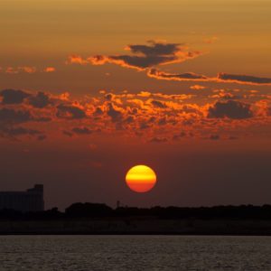 Sonnenuntergang. Eine tiefstehende Sonne stellt sich schon in geld und orange dar, beleuchtet von hinten/unten noch ein paar kleine Schönwetterwolken. Als Silhouette am Horizont sind bauten, mglw. Industriegelände zu erkennen.