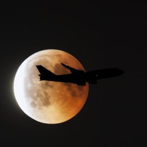 Nachtfoto: Großaufnahme vom Vollmond bzw. Blutmond, er changiert zwischen leuchtend weiß und orangerot. Vor dem Mond zieht als Silhouette ein Flugzeug vorbei, zu erkennen ist nur der Umriss und zwei Positionsleuchten