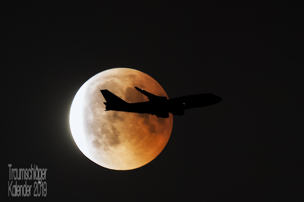 Nachtfoto: Großaufnahme vom Vollmond bzw. Blutmond, er changiert zwischen leuchtend weiß und orangerot. Vor dem Mond zieht als Silhouette ein Flugzeug vorbei, zu erkennen ist nur der Umriss und zwei Positionsleuchten