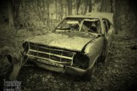 Foto nachbearbeitet auf Sepia-Effekt. Gegenstand ist ein Autowrack. Ein mindestens "Youngtimer", wahrscheinlich inzwischen schon Oldtimer. Auf dem verbeulten Kühlergrill ist gerade noch "Opel" zu erkennen, wahrscheinlich ein Kadett C. Die Scheinwerfer fehlen, ebenso die Windschutzscheibe und andere Fenster. Motorhaube, Kühlergrill und Fahrertür sind verbeult und hängen teils nur lose in den Scharnieren. Die Kofferraumklappe steht offen. Dieses Autowrack steht etwas rätselhaft mitten im Wald, fernab von Straßen und Wegen einfach auf Laub, und Laub liegt auch auf der Motorhaube.