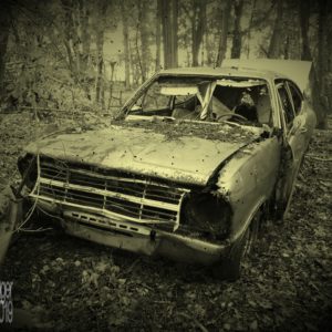 Foto nachbearbeitet auf Sepia-Effekt. Gegenstand ist ein Autowrack. Ein mindestens "Youngtimer", wahrscheinlich inzwischen schon Oldtimer. Auf dem verbeulten Kühlergrill ist gerade noch "Opel" zu erkennen, wahrscheinlich ein Kadett C. Die Scheinwerfer fehlen, ebenso die Windschutzscheibe und andere Fenster. Motorhaube, Kühlergrill und Fahrertür sind verbeult und hängen teils nur lose in den Scharnieren. Die Kofferraumklappe steht offen. Dieses Autowrack steht etwas rätselhaft mitten im Wald, fernab von Straßen und Wegen einfach auf Laub, und Laub liegt auch auf der Motorhaube.