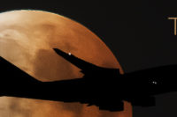 Nachtfoto: Großaufnahme vom Vollmond bzw. Blutmond, er changiert zwischen leuchtend weiß und orangerot. Vor dem Mond zieht als Silhouette ein Flugzeug vorbei, zu erkennen ist nur der Umriss und zwei Positionsleuchten