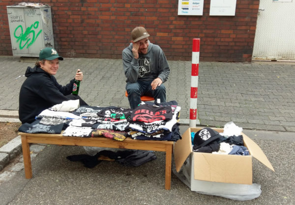 Zwei gut gelaunte Männer, davor ein niedriger Tisch und ein Karton mit diversen T-Shirts von Punk-Bands