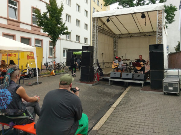 Gäste die zur Bühne schauen, darauf zwei Musiker mit Gitarren, einer singt.