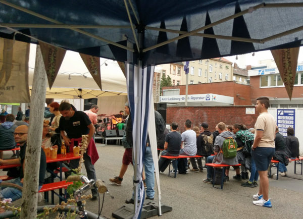 von aus einem Stand fotografiert das laufende Fest, Menschen gehen durchs Bild, viele sitzen auf Bierbänken vor der Bühne, auf der der Singer-Songwriter Yohazid auftritt