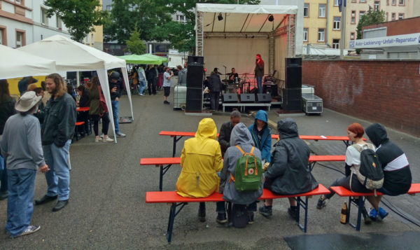 Auf der Bühne stimmen zwei Musiker den Sound ab, Festbesucher sitzen teils unter Pavillons, teils mit Kapuzen auf den Bänken vor der Bühne, da es grade mal wieder nieselt