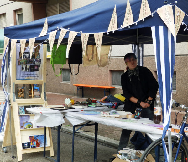 Der Stand des Traumschläger Kollektivs, ein blauer Pavillon mit zwei Tischen und einem großen Holzaufsteller mit Poster, Postkarten und Fotos, auf den Tischen Info-Materialien und die Preise der Tombola