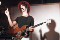 Foto von Frank Zappa. Z. steht im Licht vermutlich eines Scheinwerfers, hat eine E-Gitarre um. Sein Blick geht nach links und er hat den rechten Zeigefinger erhoben.