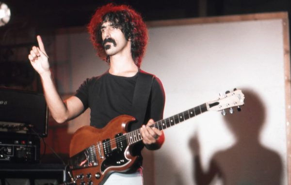 Foto von Frank Zappa. Z. steht im Licht vermutlich eines Scheinwerfers, hat eine E-Gitarre um. Sein Blick geht nach links und er hat den rechten Zeigefinger erhoben.