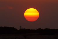 Sonnenuntergang. Eine tiefstehende Sonne stellt sich schon in geld und orange dar, beleuchtet von hinten/unten noch ein paar kleine Schönwetterwolken. Als Silhouette am Horizont sind bauten, mglw. Industriegelände zu erkennen.