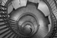 Blick von oben in ein Treppenhaus. Duchmesser ca. 10 Meter, das ganze Treppenhaus ist rund und v.a. das Treppengeländer läuft als Spirale vom Betrachter weg. Höhe 4-5 Stockwerke