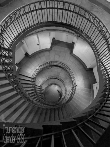 Blick von oben in ein Treppenhaus. Duchmesser ca. 10 Meter, das ganze Treppenhaus ist rund und v.a. das Treppengeländer läuft als Spirale vom Betrachter weg. Höhe 4-5 Stockwerke