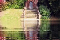 Ein gemauertes Portal aus rotem Standstein, hinauf führen 7 Treppenstufen. Um das Portal herum ein grüner und viele rotblättrige Büsche und Bäume. Auf der Ebene, wo der Fotograf steht ist das Bild bearbeitet, statt des Originalgrundes wurde eine Spiegelung des oberen Teils eingefügt, also von Treppe, Portal und Bäumen, wie in einem Weiher.