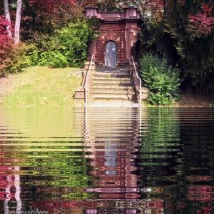 Ein gemauertes Portal aus rotem Standstein, hinauf führen 7 Treppenstufen. Um das Portal herum ein grüner und viele rotblättrige Büsche und Bäume. Auf der Ebene, wo der Fotograf steht ist das Bild bearbeitet, statt des Originalgrundes wurde eine Spiegelung des oberen Teils eingefügt, also von Treppe, Portal und Bäumen, wie in einem Weiher.