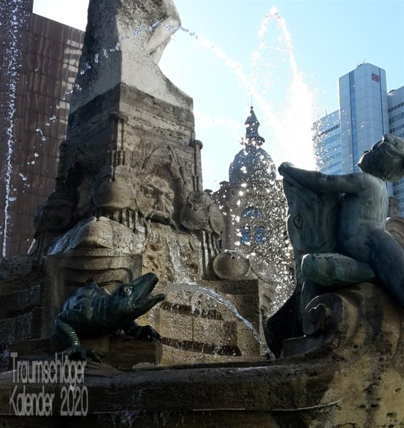 Foto eines Brunnens im Gegenlicht. Verschiedene Wasserspeier, ein Wels, ein Putto, ein Titan, steuern Fontänen bei. Die Fontänen sind mit jedem Tropfen zu erkennen, nicht verwischt als ein Strahl. Im Hintergrund noch zwei Hochhausspitzen.