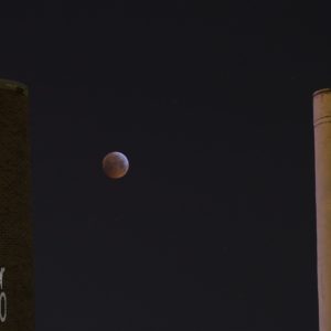 Nachtfoto: Zwei Schornsteine eines Heizkraftwerks, vom städtichen Lichtkonzept leicht angestrahlt. Zwischen den Schornsteinen nicht ganz mittig ein Vollmond in dunkelroter Färbung, vom Blutmond oder ener (partiellen) Mondfinsternis. Einige Betrachter sagten spontan: "Mond-Pong!"