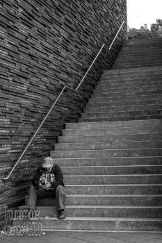 Schwarzweiß-Foto. Auf den untersten drei Stufen einer langen, in drei Abschnitte gegliederten Treppe, sitzt ein Mann, mit Baseballcap und Fanschal, er schaut auf sein Handy. Den Rest des Bildes dominiert die Treppe und die sie begrenzende Wand, die aus rauhen Steinen grob zusammengesetzt ist, mit viel Struktur.
