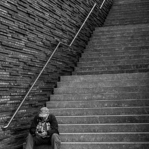 Schwarzweiß-Foto. Auf den untersten drei Stufen einer langen, in drei Abschnitte gegliederten Treppe, sitzt ein Mann, mit Baseballcap und Fanschal, er schaut auf sein Handy. Den Rest des Bildes dominiert die Treppe und die sie begrenzende Wand, die aus rauhen Steinen grob zusammengesetzt ist, mit viel Struktur.