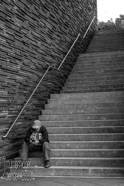 Schwarzweiß-Foto. Auf den untersten drei Stufen einer langen, in drei Abschnitte gegliederten Treppe, sitzt ein Mann, mit Baseballcap und Fanschal, er schaut auf sein Handy. Den Rest des Bildes dominiert die Treppe und die sie begrenzende Wand, die aus rauhen Steinen grob zusammengesetzt ist, mit viel Struktur.