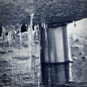 Detailfoto eines Brunnens. Im Hintergrund ein Wasserrohr, im Vordergrund herunterlaufendes und heruntertropfendes Wasser, das von der Unterseite eines abgerundeten Steins heruntertropft.