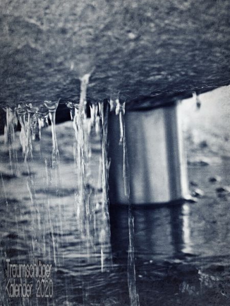 Detailfoto eines Brunnens. Im Hintergrund ein Wasserrohr, im Vordergrund herunterlaufendes und heruntertropfendes Wasser, das von der Unterseite eines abgerundeten Steins heruntertropft.