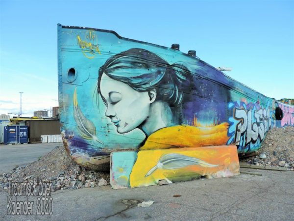 Dokumentarisches Foto: Graffito einer jungen Frau in Türkistönen, die Haare zum Pferdeschwanz gebunden und mit geschlossenen Augen leicht lächelnd, Malgrund für das Graffito ist ein auf Land gelegtes Schiff, vermutlich ein Fluss-Lastkahn. Graffitto von Hanasaari Hanaholmen in Helsinki, Finnland