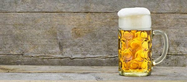 Ein mit hellgelbem Bier gefüllter Maßkrug. Der Schaum fält ganz leicht über den Rand des Glases, rinnt aber nicht herunter. Tisch und Hintergrund sind altes, vom Wetter patiniertes grau gewordenes Holz. Sonst nichts.