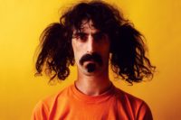 Frank Zappa. vor gelbem Hintergrund, mit orangenem T-Shirt (vor gelb fast unsichtbar).Mit Schnurrbart, Goatee und links und rechts je einem zerzausten "Pferdeschwanz" (im Gegensatz zum geflochtenen Zopf)