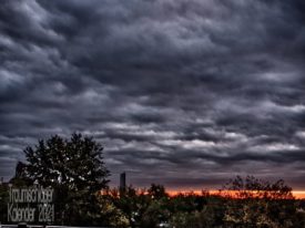 Aufnahme eines gewittrigen Himmels. Land/-schaft ist nur im unteren Viertel des BIldes zu sehen, vor allem ein Baum und wenige Büsche. am Horizont bricht kräftiges Abendrot durch die Wolken, die den Rest des BIldes beherrschen. Sie sind unterschiedlich dunkel grau, vom Wind vorangetrieben, zerfestzt. Die Aufnahme ist mit HDR-Technik verstärkt um die Theatralik zu betonen