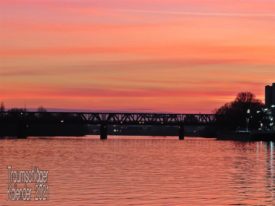 In kräftigem Abendrot und Gegenlicht eine Metallfachwerkbrücke über einen Fluß. Durchs Gegenlicht alles nur als Silihoutte zu sehen, an den Seiten des BIldes ist noch die Uferbewachsung zu erkennen