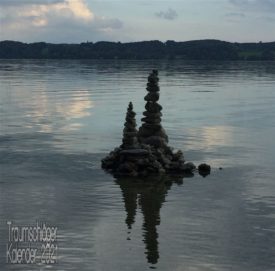 im Uferbereich eines Sees hat jemand Steine aufgetürmt. Stück für Stück aufeinander gesetzt und ausbalanciert ist eine Skulptur entstanden, ca 1m hoch und mit zwei hohen, schlanken Türmen. Im Bildhintergrund ist das andere Ufer zu sehen